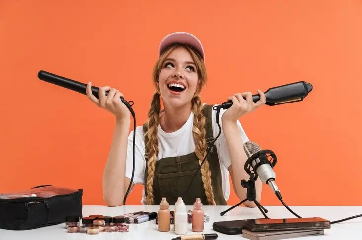 Young girl holding hair straighteners while recording blog broadcast about new cosmetic products isolated on orange wall